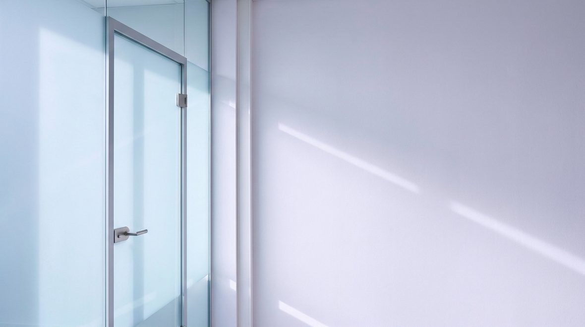 A glass office partition catching cool daylight, with soft diagonal shadow gradients falling across a pale wall.